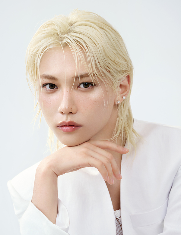 밝은 금발 머리에 흰 재킷을 입은 모델이 턱을 손으로 받치고 은은한 미소를 짓고 있으며, 헤라의 광채 피부 표현을 자연스럽게 보여줍니다.